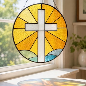 May include: Round stained glass window with a white cross in the center. The window features a yellow and orange sunburst design, with blue and green accents at the bottom. It is hanging from a black chain.