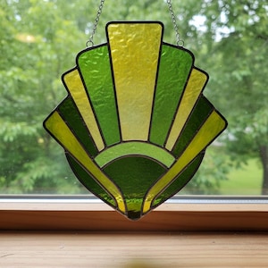 May include: A stained glass suncatcher in shades of green and yellow, with a fan-like design. The glass is segmented with black lines and hangs from a chain. The suncatcher is displayed in front of a window.