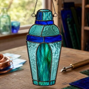 May include: A stained glass cocktail shaker suncatcher featuring a geometric design with textured light blue, teal, and deep cobalt blue glass panels. The piece is constructed with dark metal solder lines and includes a metal chain for hanging.