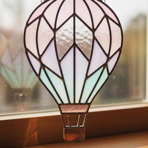 Könnte beinhalten: Ein Glasfenster-Sonnenfänger in Form eines Heißluftballons. Der Ballon hat ein geometrisches Design mit rosa, weißen und türkisfarbenen Glasscheiben, die von dunklen Linien umrahmt sind. Ein kleiner brauner Korb hängt darunter.
