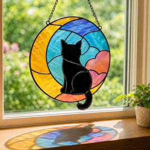 May include: Stained glass suncatcher featuring a black cat silhouette perched on a crescent moon. The moon is yellow, with a blue and turquoise sky, and pink and orange clouds. The suncatcher hangs from a black chain.