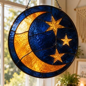 Op de afbeelding: Een ronde glas-in-lood raamdecoratie met een hemels ontwerp. Het heeft een gele maan en drie gouden sterren tegen een blauwe glazen achtergrond. Het raam is zwart omlijst en hangt aan een ketting.