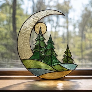 Puede incluir: Un atrapasoles de vitral en forma de media luna con una luna texturizada de color crema, un pequeño sol amarillo y tres pinos verdes sobre un paisaje de colinas verdes y un elemento de agua azul claro con bordes de metal oscuro.