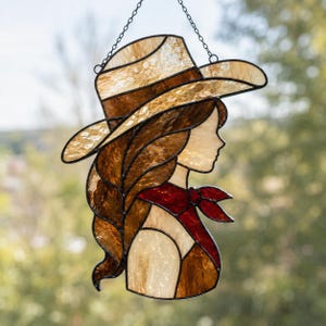 Könnte beinhalten: Ein Buntglas-Suncatcher, der das Profil einer Person mit einem beigen Hut mit breiter Krempe und einem roten Halstuch zeigt. Das Stück besteht aus strukturierten bernsteinfarbenen und braunen Glasteilen, die mit dunklen Metallnähten verbunden sind.