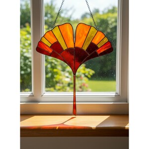 Op de afbeelding: Een gebrandschilderd glazen ginkgo blad zonnevanger in tinten rood, oranje en geel hangt voor een raam. De zonnevanger wordt opgehangen door een ketting en werpt een schaduw op de houten vensterbank. De achtergrond toont een groene, bladerrijke buitenscène.