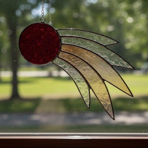 May include: Stained glass suncatcher featuring a red circle and layered, curved wings in green, gold, and yellow. The suncatcher hangs from a silver chain. The background is a blurred view of a green, outdoor scene.