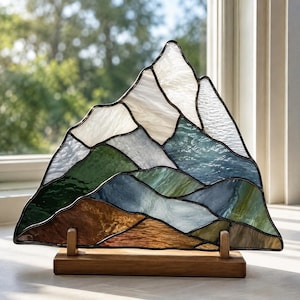 May include: A triangular stained glass panel depicting a mountain landscape with white, blue, green, and brown textured glass segments joined by dark metal solder lines, supported by a rectangular wooden base with two upright wooden supports.