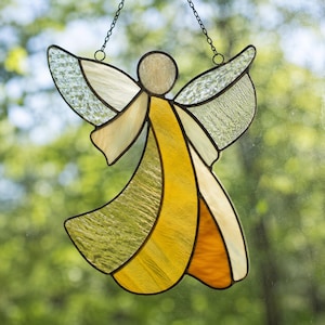 May include: A stained glass angel suncatcher featuring textured clear glass wings and a body composed of yellow, amber, and cream-colored glass panels. The figure is outlined in dark metal solder and hangs from a thin black metal chain.
