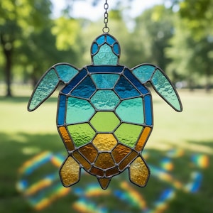 May include: Stained glass turtle suncatcher with a chain for hanging. The turtle features a mosaic design with shades of blue, green, and amber. The suncatcher is suspended against a blurred green background, suggesting an outdoor setting.