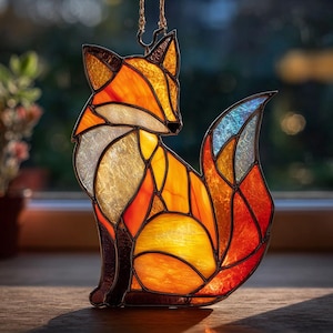 Puede incluir: Atrapasoles de zorro de vidriera en tonos naranja, amarillo y azul. El zorro está sentado con un diseño de mosaico detallado. Se adjunta una cadena dorada para colgarlo. La obra de arte se exhibe sobre una superficie de madera.