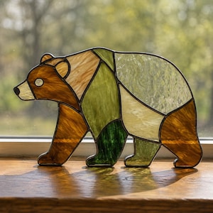 Op de afbeelding: Een glas-in-lood beren-zonnevanger gemaakt van diverse stukken getextureerd glas in tinten bruin, bosgroen, olijf en crème. De beer is afgebeeld in een wandelend profiel, waarbij donkere metalen soldeerlijnen elk glassegment omlijnen.