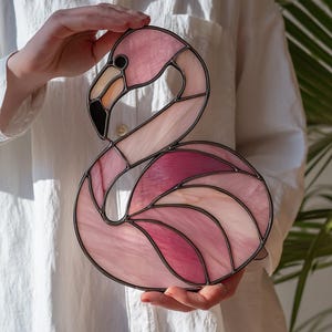 May include: A stained glass flamingo sculpture in shades of pink, with a black beak and a dark metal frame. The flamingo is held against a white shirt and a green leafy background.