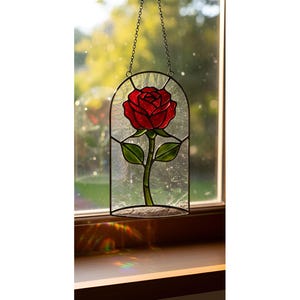 Peut inclure: Panneau en vitrail représentant une rose rouge avec des feuilles et une tige vertes. La rose est enfermée dans une arche en verre clair, suspendue par une chaîne. Le panneau est exposé devant une fenêtre.