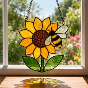 May include: Stained glass sunflower with a bee, featuring yellow petals, a brown center, and green leaves. The bee has black and yellow stripes and translucent wings. The artwork hangs from a black chain, perfect for window decor.