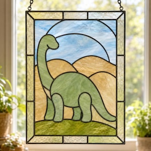 May include: A rectangular stained glass panel featuring a long-necked green dinosaur standing on a grassy hill against a backdrop of tan rolling hills and a light blue sky. The piece is framed by a border of textured clear glass segments.