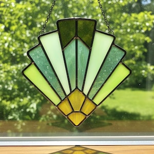 May include: A stained glass suncatcher in a fan design, featuring shades of green and yellow. The geometric pattern is framed with dark lines and suspended by a chain. The suncatcher is displayed in front of a blurred green background.