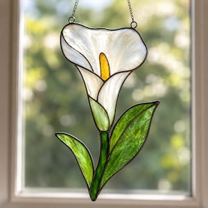 May include: A stained glass suncatcher shaped like a calla lily flower. The piece features a large, translucent white petal with a central yellow spadix, supported by a green stem and two green leaves, all outlined in dark metal solder.