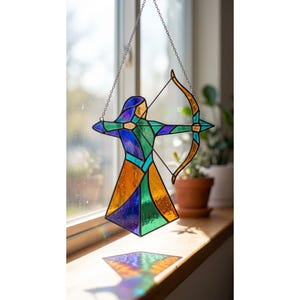 Peut inclure: Suncatcher en vitrail représentant un archer avec un arc et une flèche. L'archer est composé de morceaux de verre bleus, verts et oranges, suspendus à une chaîne argentée. Le capteur solaire est exposé devant une fenêtre.