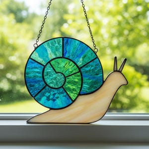 May include: Stained glass snail suncatcher with a spiral shell in shades of blue and green. The snail's body is a light beige colour, and it hangs from a chain. Decorative art for windows.