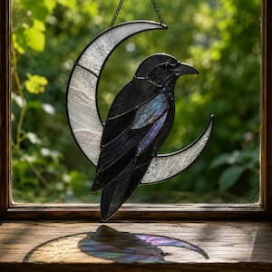 May include: A stained glass suncatcher featuring a black crow perched on a crescent moon. The moon is made of textured white glass, while the crow consists of dark, iridescent glass pieces held together by black metal solder lines.