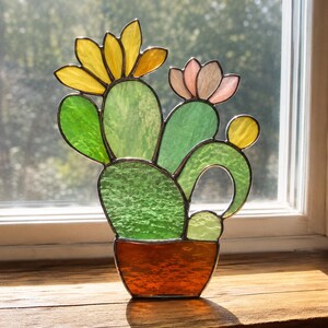 May include: A stained glass cactus ornament featuring green textured glass pads in a brown pot. The cactus has a large yellow flower on the left and a smaller pale pink flower on the right, with a small yellow circular accent on the far right side.