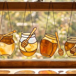 Pode incluir: Quatro peças decorativas de janela em vitral com potes de mel e colheres de mel de madeira. Os potes têm várias formas e tamanhos, com tons de âmbar, amarelo e castanho. Cada pote é suspenso por uma corrente.