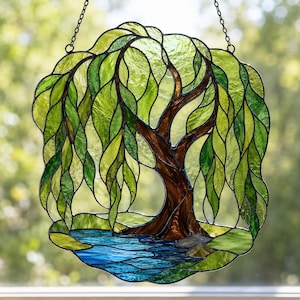 Op de afbeelding: Een glas-in-lood suncatcher met een treurwilg met gestructureerde groene glazen bladeren en een bruine glazen stam. De boom is geplaatst boven een stuk blauw glas dat water voorstelt, alles bijeengehouden door donkere loden lijnen.