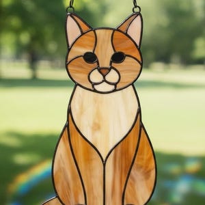 May include: Stained glass cat suncatcher with a chain for hanging. The cat is depicted in shades of brown and tan with black accents. The cat is sitting upright with its tail curled to the side. The background is blurred green.