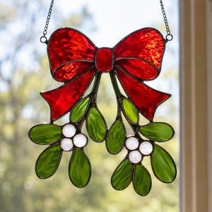 May include: A stained glass suncatcher featuring a large, textured red bow at the top with two sprigs of green mistletoe leaves and clusters of small, opaque white berries hanging below, all held together by dark metal solder and a hanging chain.