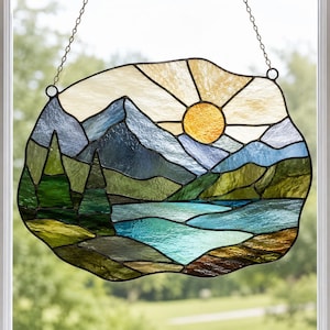 May include: A stained glass suncatcher featuring a landscape scene with a bright yellow sun, blue mountain peaks, green pine trees, and a turquoise lake. The piece is framed in dark metal solder lines and hangs from a thin silver-colored metal chain.