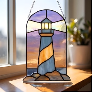 May include: Stained glass lighthouse with a blue and yellow striped tower, set against a sunset sky of purple, orange, and yellow. The lighthouse is framed with a black border and hangs from a chain. The base of the lighthouse rests on a rocky shoreline.