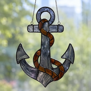 May include: A stained glass anchor suncatcher featuring textured grey glass panels and a coiled amber-coloured rope detail. The anchor is framed in dark metal solder lines and hangs from a thin metal chain, designed for display in a window.