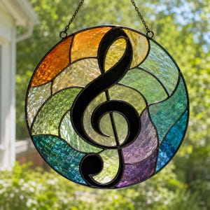 May include: A circular stained glass suncatcher featuring a black metal treble clef centred over a multi-coloured background. The glass segments display a gradient of orange, yellow, green, blue, and purple hues with a textured, rippled finish.