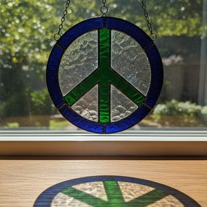 Könnte beinhalten: Ein kreisförmiges Peace-Zeichen aus Glas mit blauem Rand und grünem Peace-Zeichen. Es hängt an einer Kette. Das Symbol wirft einen Schatten auf eine Holzoberfläche vor einem Fenster.