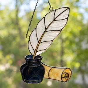 May include: A stained glass suncatcher featuring a white feather quill standing upright in a dark blue inkwell, with a rolled parchment scroll in light yellow attached to the side. The piece is suspended by a thin black metal chain.