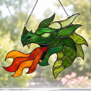 Può includere: Un acchiappasole in vetro colorato a forma di testa di drago di profilo, caratterizzato da varie tonalità di vetro verde con linee di saldatura in metallo scuro. Il drago è raffigurato mentre sputa fiamme in vetro arancione e rosso vivo.