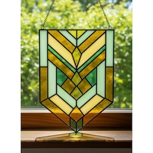 May include: Stained glass panel with a geometric design. The panel features a combination of yellow, green, and clear glass pieces, framed with black lines. It is suspended by a chain and has a matching base for display.