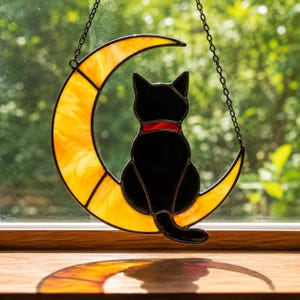Op de afbeelding: Een zwarte kat met een rode halsband zit op een gele glas-in-lood maan. De zonnevanger hangt aan een ketting, met een natuurlijke groene achtergrond zichtbaar door het raam.