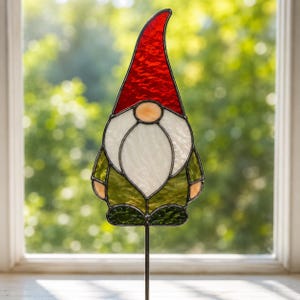 May include: A stained glass garden stake shaped like a gnome. The gnome features a tall, textured red hat, a pale peach-coloured nose, a large white beard, and a dark green tunic. The pieces are joined by dark metal solder lines on a thin rod.