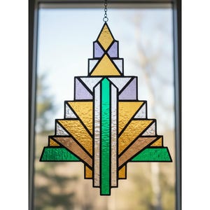 May include: A geometric stained glass suncatcher. The design incorporates gold, green, purple, and brown glass, framed by black lines. It's suspended by a chain, suitable for window decoration. The artwork is symmetrical.