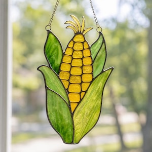 May include: A stained glass suncatcher shaped like an ear of corn with bright yellow kernels and textured green husks. The piece is constructed with dark metal solder lines and includes a thin metal chain attached to the top for hanging display.