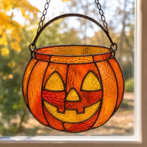 Könnte beinhalten: Ein Sonnenfänger aus Buntglas in Form eines Kürbiseimers mit orangefarbenen und gelben, strukturierten Glasscheiben, die durch dunkle Metalllötlinien verbunden sind. Das Gesicht hat dreieckige Augen, eine kleine Nase und ein breites Grinsen.
