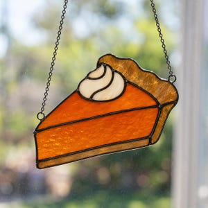 Könnte beinhalten: Ein Buntglas-Suncatcher in Form eines Stücks Kürbiskuchen mit einem Klecks Schlagsahne obenauf. Das Stück besteht aus strukturiertem orangefarbenem Glas für die Füllung, beigefarbenem Glas für den Rand und undurchsichtigem weißem Glas für die Sahne, alles zusammengehalten durch schwarze Metalllötlinien und aufgehängt an einer dünnen Metallkette.