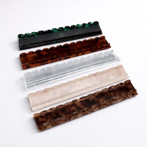 May include: A collection of five rectangular hair barrettes in various colors and patterns. The barrettes have a scalloped edge and are in shades of green, brown, clear, pink, and tortoiseshell. The barrettes are made of a shiny material.