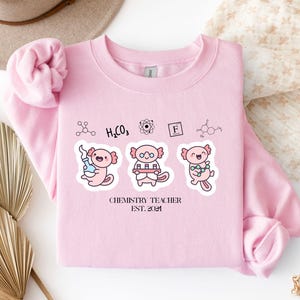 Könnte beinhalten: Ein hellrosa Sweatshirt mit Rundhalsausschnitt und einer Grafik von drei Cartoon-Axolotln, die Chemieexperimente durchführen, umgeben von wissenschaftlichen Symbolen und dem schwarzen Text "CHEMISTRY TEACHER EST. 2021" auf der Vorderseite.