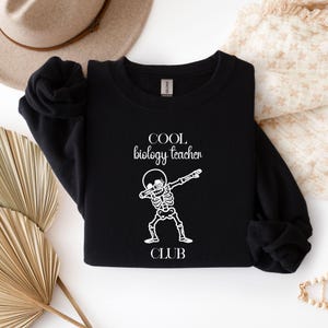 Könnte beinhalten: Ein schwarzes Langarm-Sweatshirt mit weißem Text, der COOL biology teacher CLUB lautet, und einer Grafik eines Skeletts, das eine Dab-Pose einnimmt. Das Kleidungsstück hat einen Rundhalsausschnitt und lange Ärmel mit gerippten Bündchen.