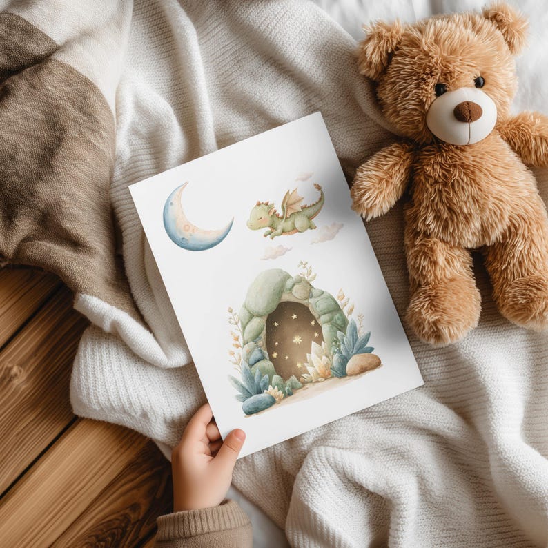 May include: A watercolor print featuring a crescent moon, a green dragon, and a cave with stars. The print is held by a child's hand and is next to a brown teddy bear on a white blanket.