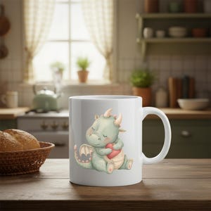 May include: White ceramic mug featuring a watercolor illustration of a light green dragon hugging a red heart. The dragon has small horns and closed eyes. The mug is on a wooden table in a kitchen setting.