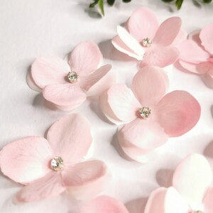 Op de afbeelding: Close-up van verschillende lichtroze stoffen bloemen met een sprankelend strass steentje in het midden. De bloemblaadjes hebben een delicate, gelaagde uitstraling. Groene bladeren zijn zichtbaar op de achtergrond. Deze bloemen zijn geschikt voor knutselen en versieringen.