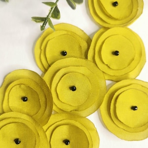 Könnte beinhalten: Nahaufnahme von mehreren gelben Stoffblumenverzierungen mit schwarzen Zentren. Die Blumen haben ein geschichtetes Blütenblattdesign. Grüne Blattzweige befinden sich in der oberen linken Ecke. Die Blumen sind auf einer weißen Oberfläche angeordnet.